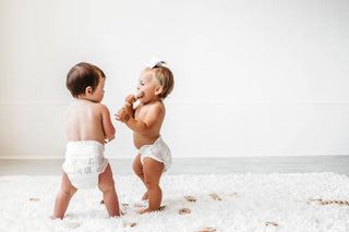 Two babies in diapers standing side by side on a white rug with a white wall background.
