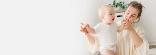 A woman in a cream-colored blouse holds a baby wearing a white long-sleeve shirt and diaper, posing against a bright white wall with a small green plant in the background.
