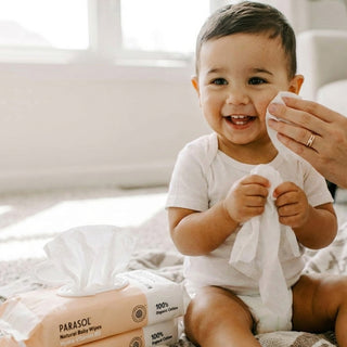 A baby sitting on a bed while an adult hand wipes their face with a wipe. Two packs of Parasol baby wipes are visible in the foreground.
