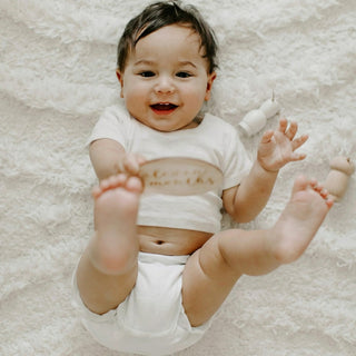 A smiling baby lying on a white textured blanket, wearing a white short-sleeve t-shirt.
