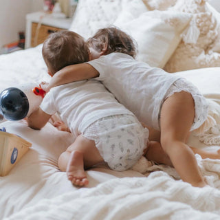 Two toddlers hugging while lying on a bed, wearing matching white t-shirts and white patterned diaper cover pants.
