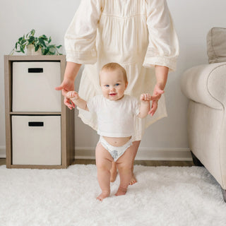 A baby wearing a white t-shirt and diaper stands on a white shag rug while holding an adult's hands. The adult is wearing a cream-colored dress.
